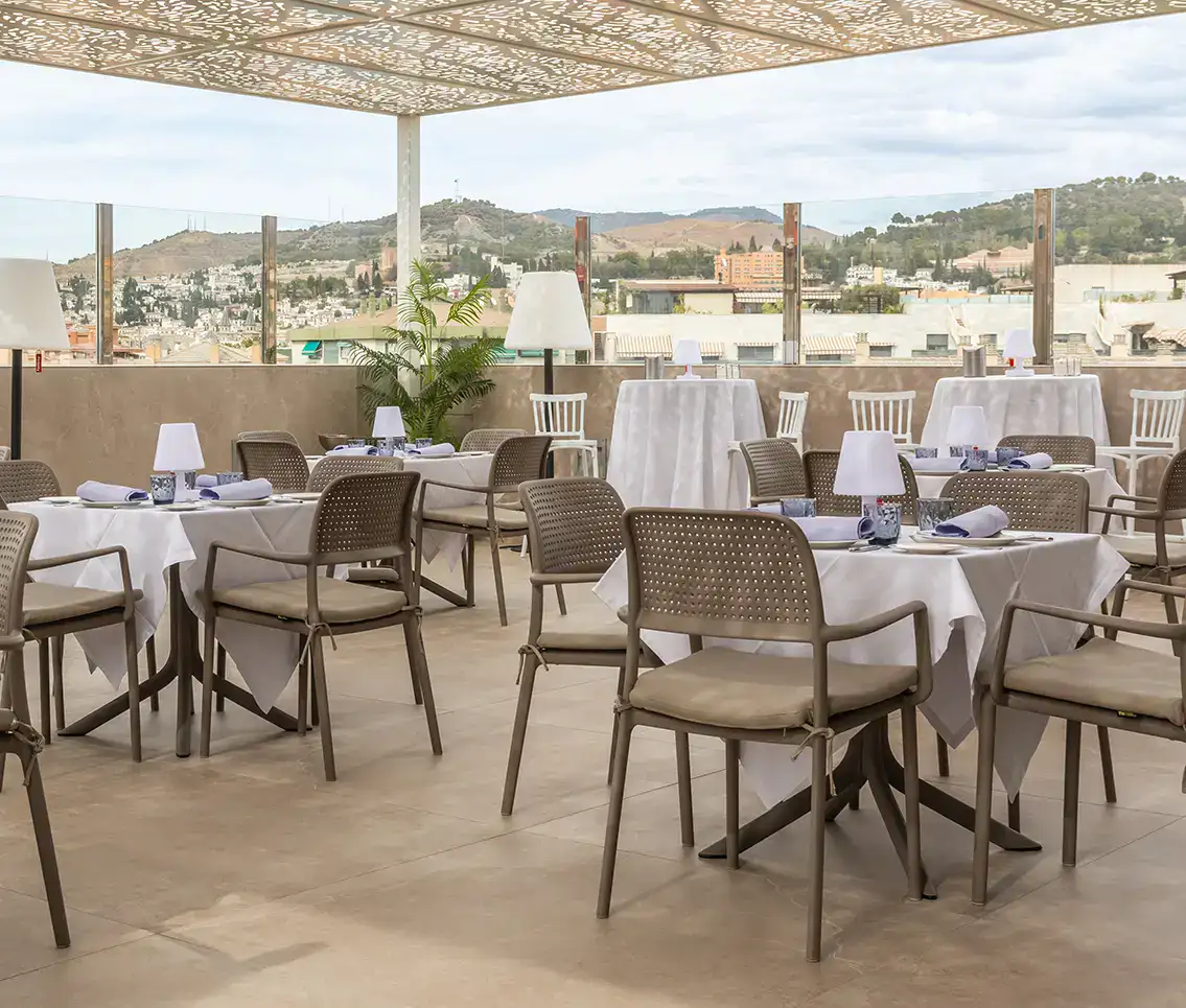 Terraza de restaurante con mesas blancas y sillas marrones; vista de colinas y ciudad.