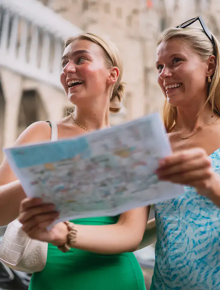 Dos mujeres sonrientes mirando un mapa tur&iacute;stico en una calle hist&oacute;rica.