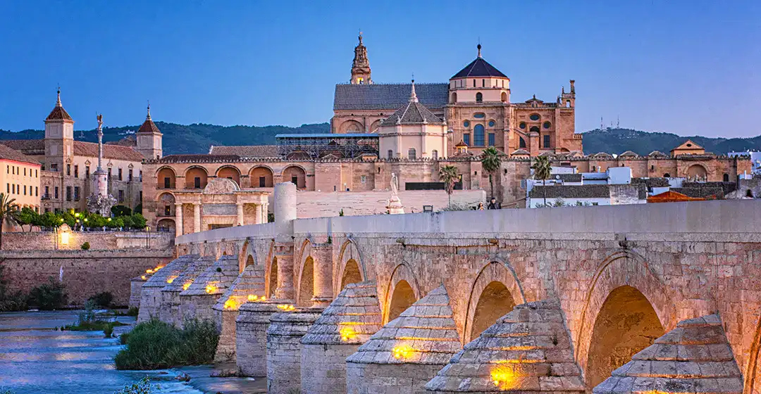 Puente de piedra iluminado al anochecer con una ciudad hist&oacute;rica al fondo.