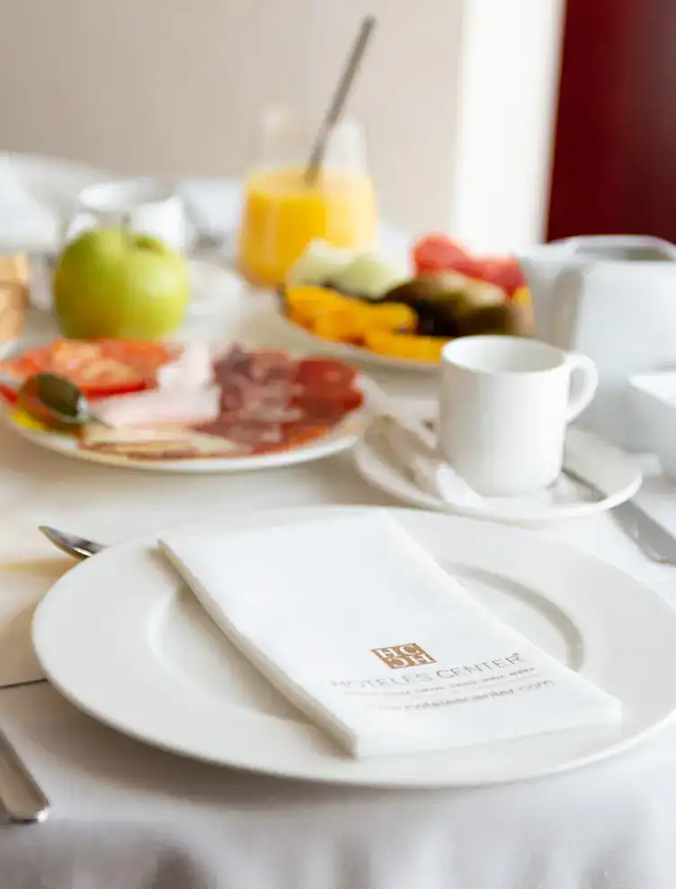 Desayuno con jugo, frutas, embutidos y taza en una mesa con mantel blanco.