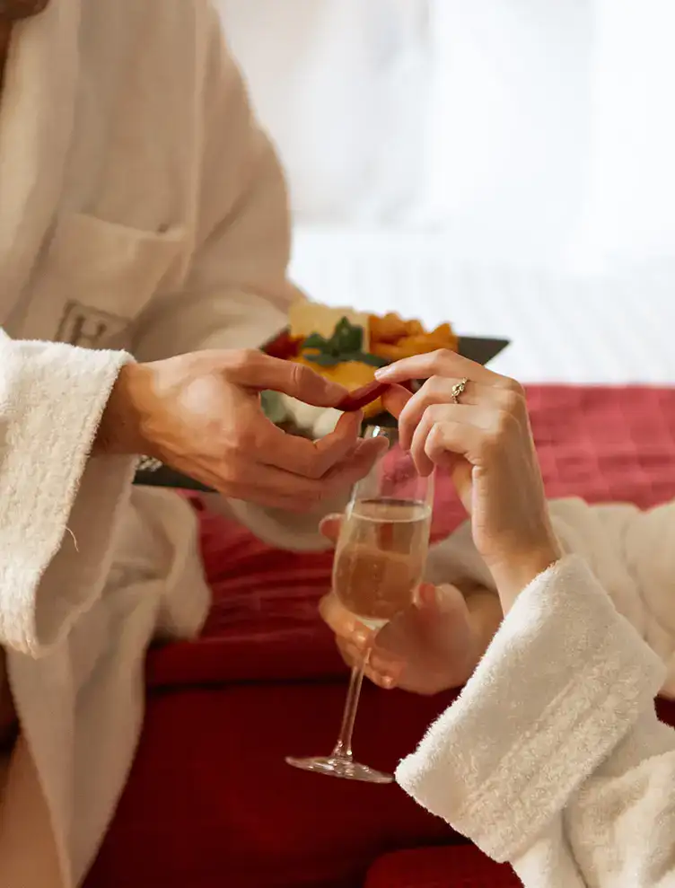 Manos brindando con champ&aacute;n y un plato de bocadillos en la cama.