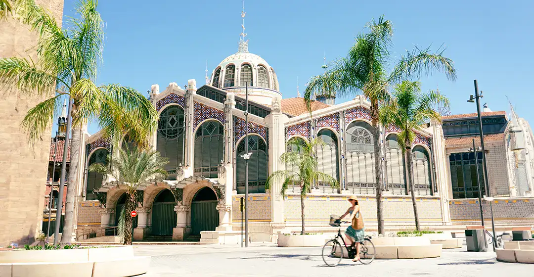 Persona en bicicleta frente a un edificio hist&oacute;rico con &aacute;rboles de palmera.