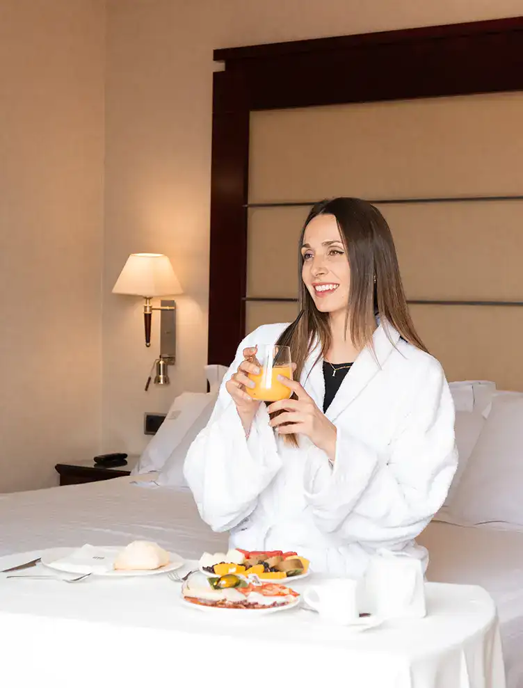 Mujer sonriente en bata blanca, disfrutando desayuno en la cama con jugo de naranja.