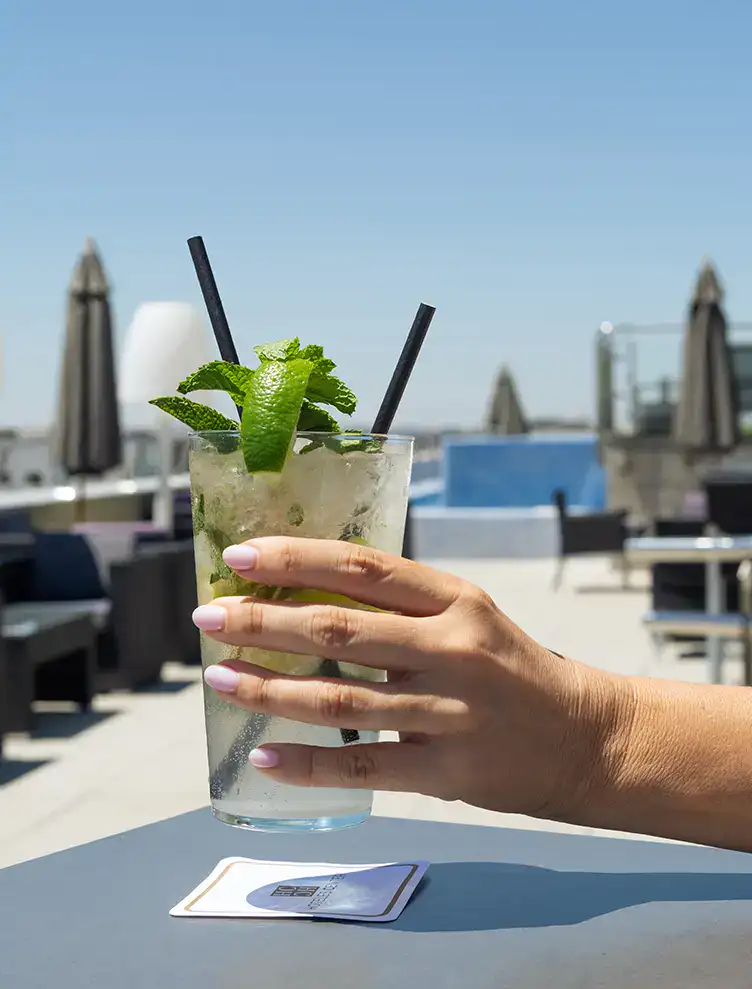 Mano sostiene un c&oacute;ctel con menta y lim&oacute;n en una terraza al aire libre.