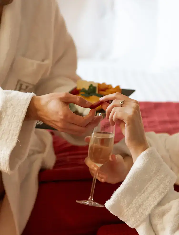 Personas en batas compartiendo bocadillos y brindando con champ&aacute;n en la cama.