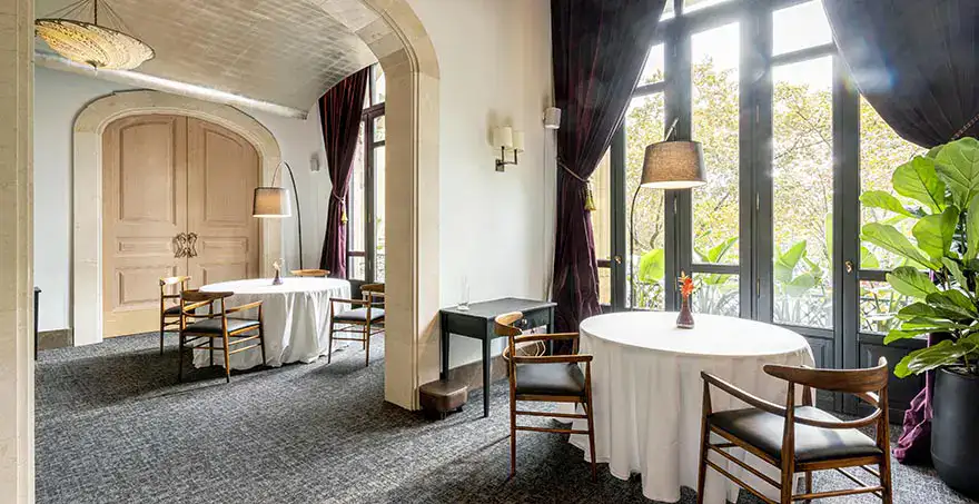 Sala elegante con mesas redondas, sillas de madera y grandes ventanales.