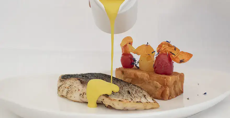 Pescado con salsa amarilla servido con guarnici&oacute;n decorada en un plato blanco.