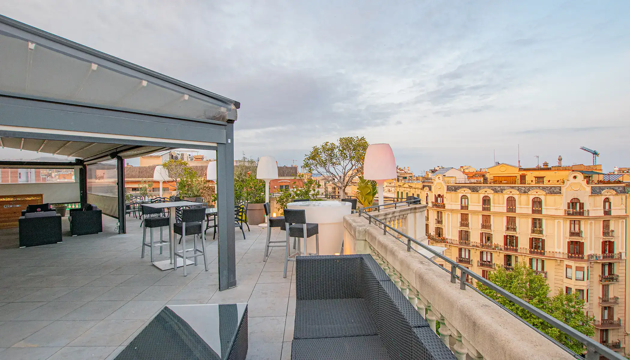 Terraza con mesas y sillas, vista de edificios y cielo parcialmente nublado.