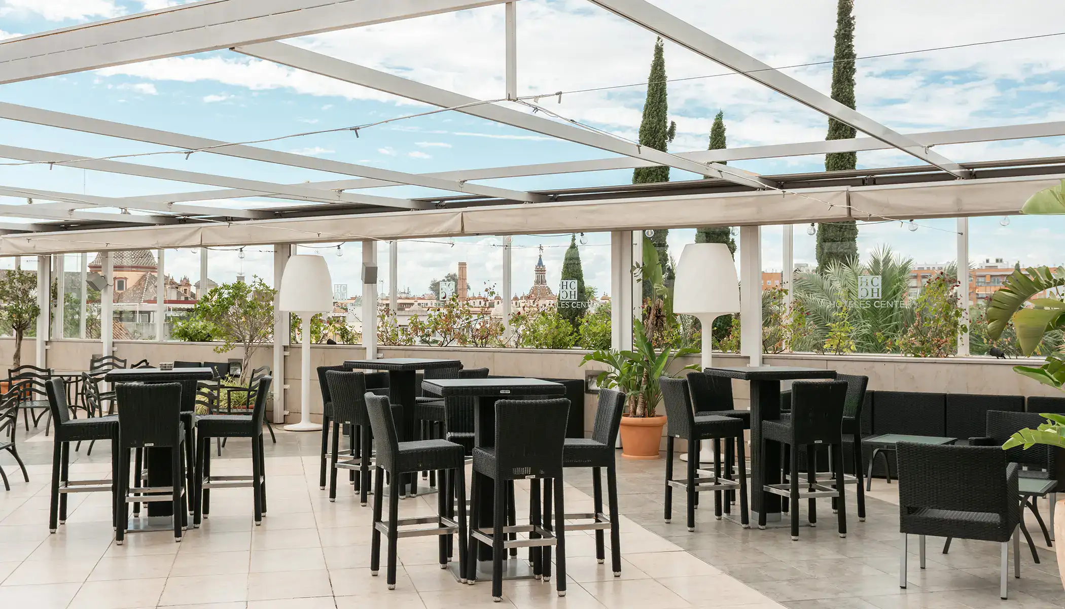Terraza con mesas y sillas negras, plantas y vista urbana de fondo.
