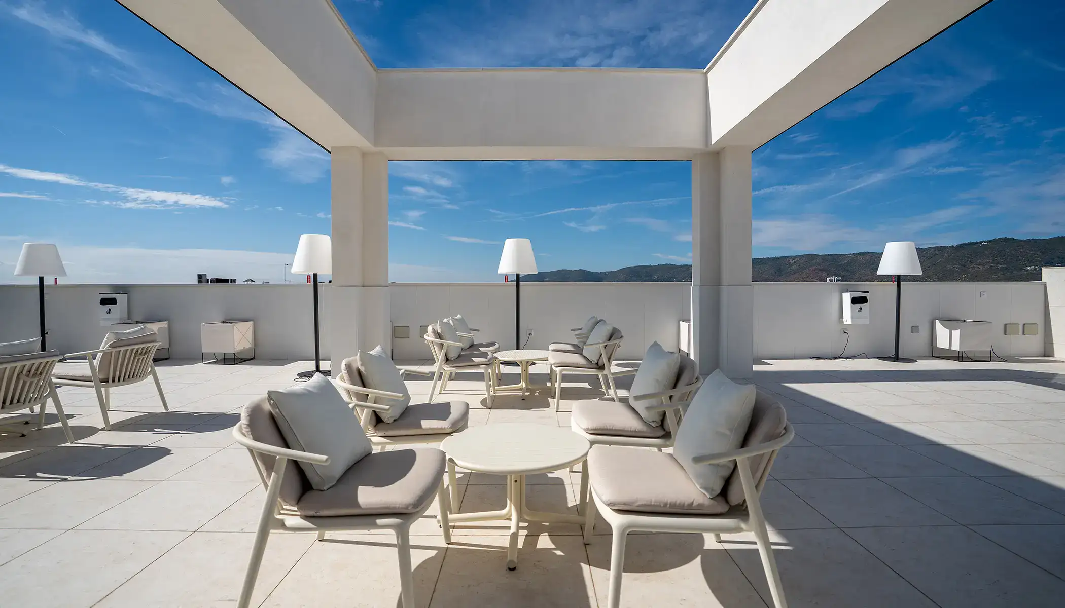 Terraza minimalista con sillas y mesas blancas bajo un cielo azul.