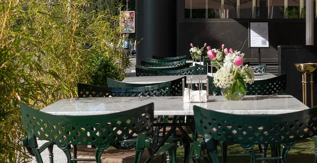 Mesas al aire libre con sillas verdes y flores en jarrones de vidrio.