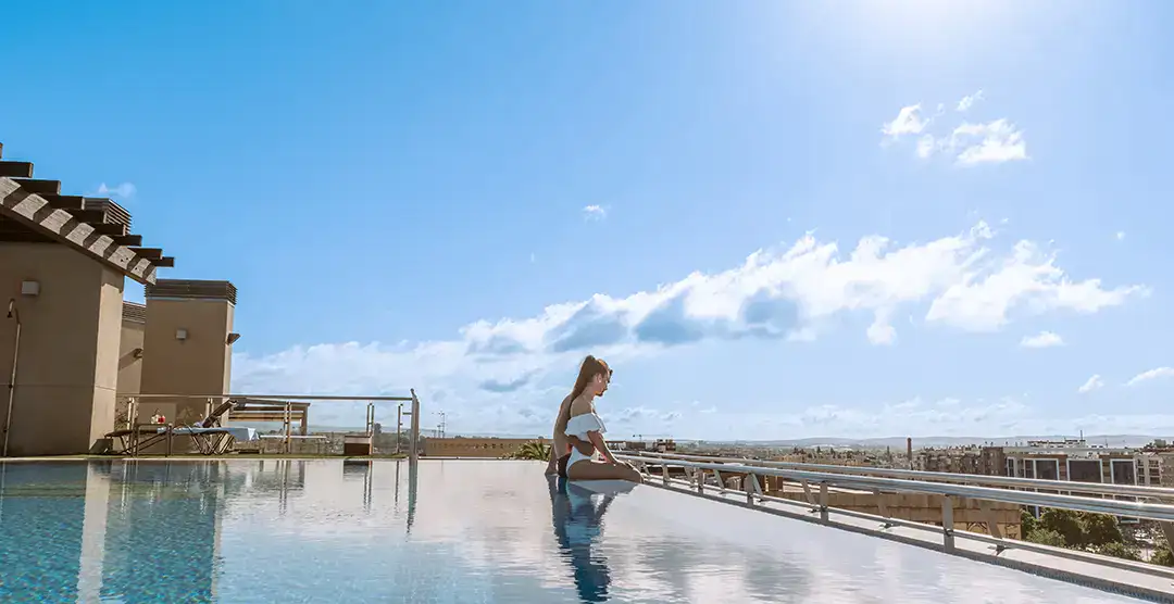 Mujer sentada al borde de una piscina en una azotea bajo un cielo despejado.
