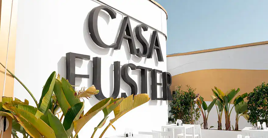 Fachada de un edificio con el letrero "Casa Fuster" y plantas alrededor.