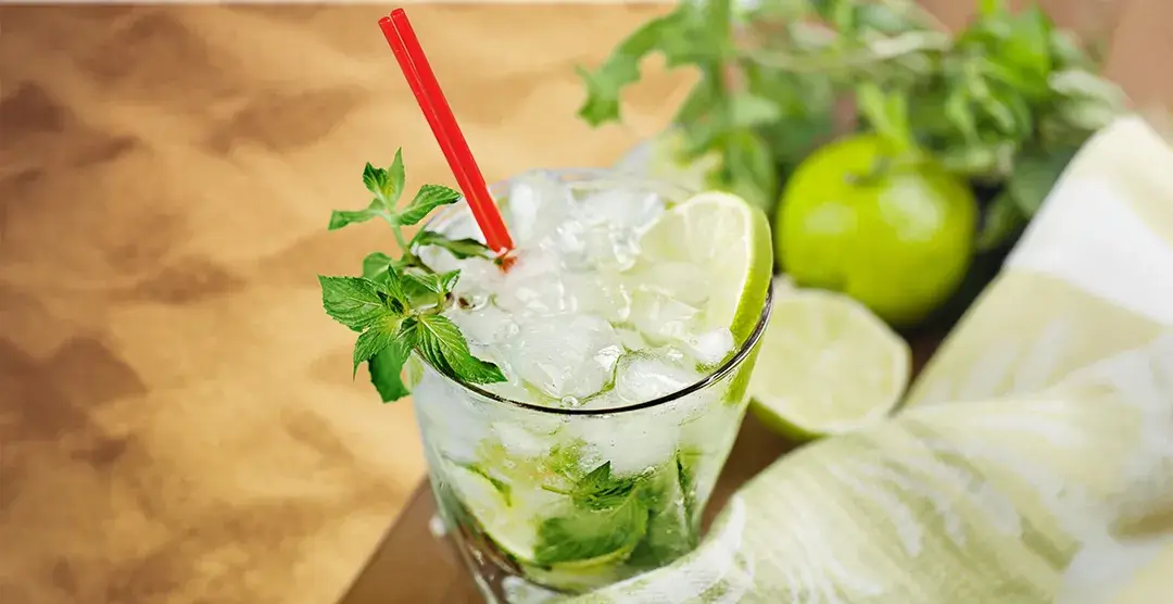 C&oacute;ctel de mojito con menta, lima y pajita roja, adornado con hojas frescas.