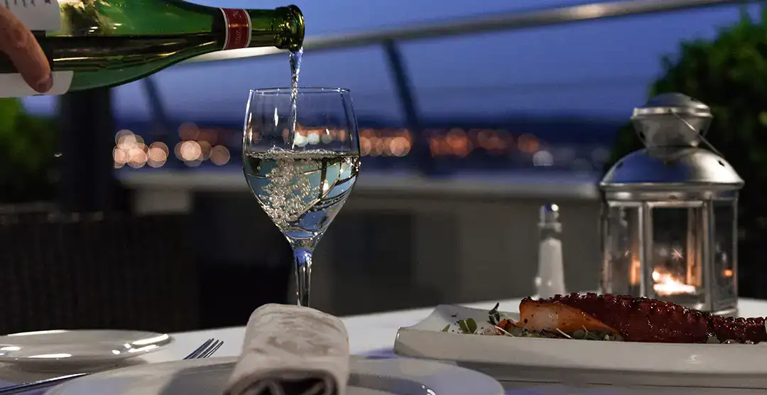 Vino sirvi&eacute;ndose en copa junto a plato de comida y l&aacute;mpara al anochecer.