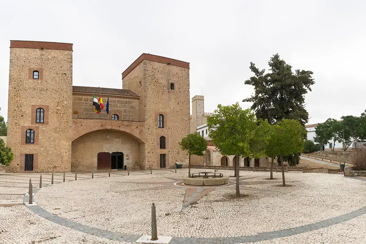 Plaza empedrada con edificio hist&oacute;rico y banderas, rodeada de &aacute;rboles.