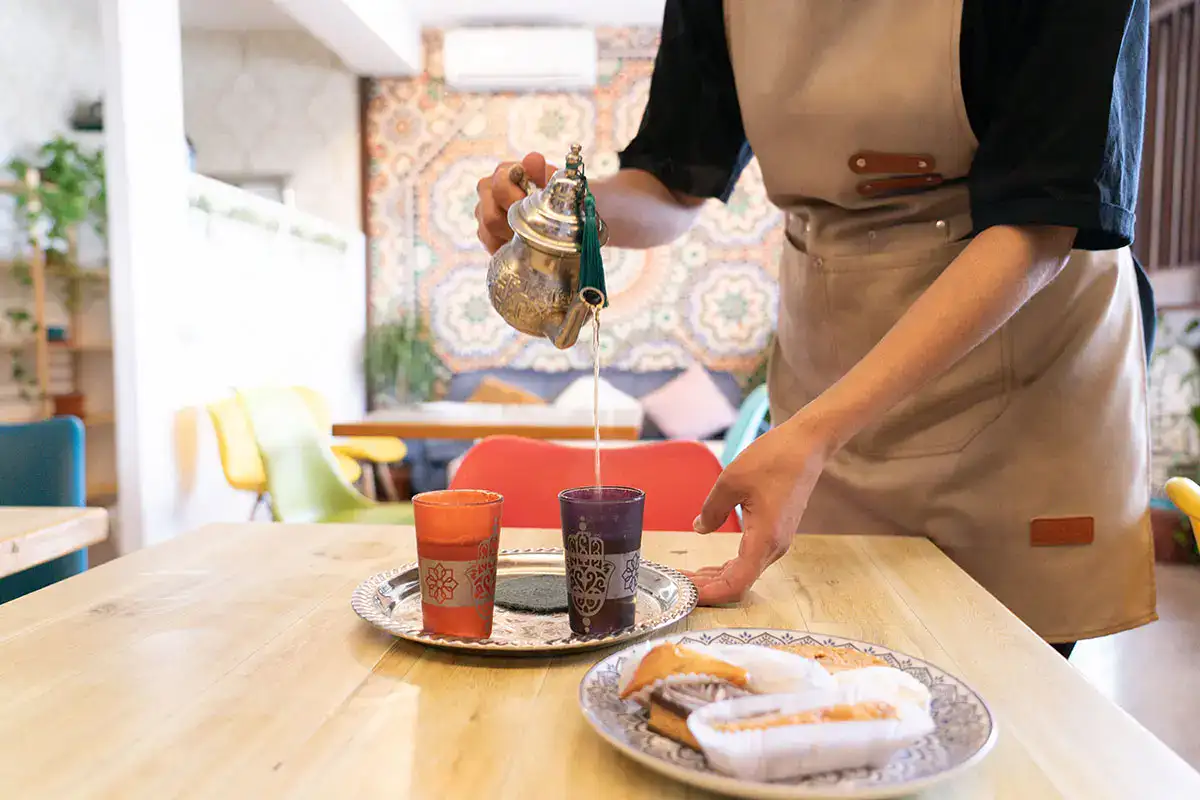 Persona sirviendo t&eacute; en vasos decorados sobre mesa con dulces y fondo colorido.
