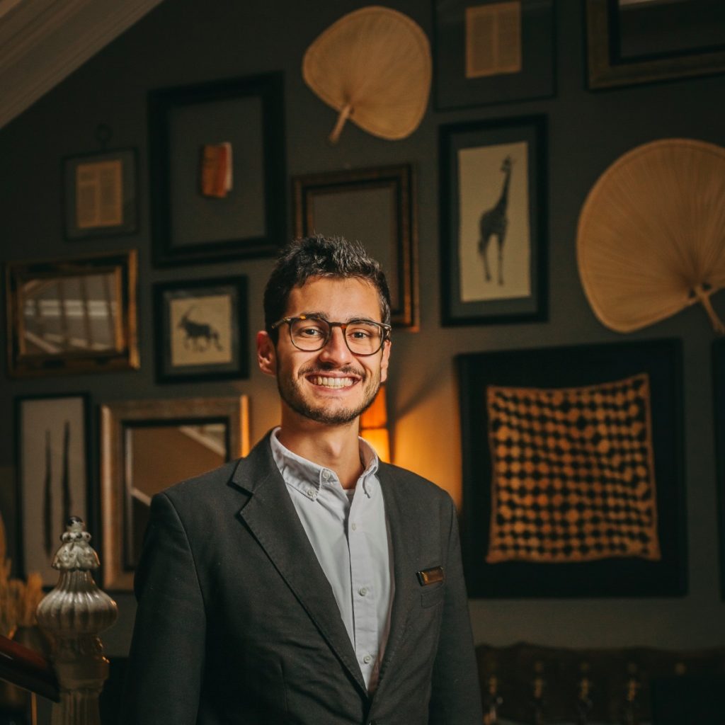 Smiling person in glasses, suit jacket; art and fans decorate dimly lit background wall.