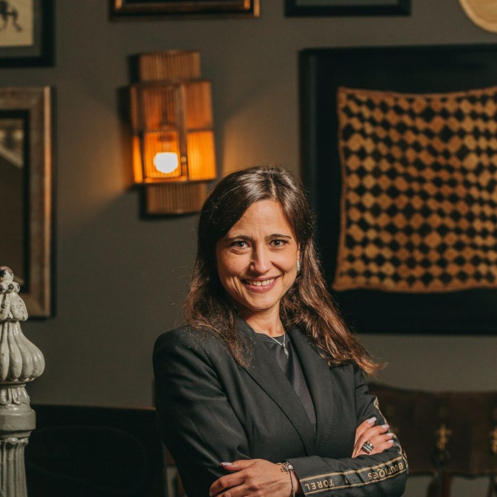 Woman in black blazer smiling, arms crossed, against patterned wall decor and warm lighting.