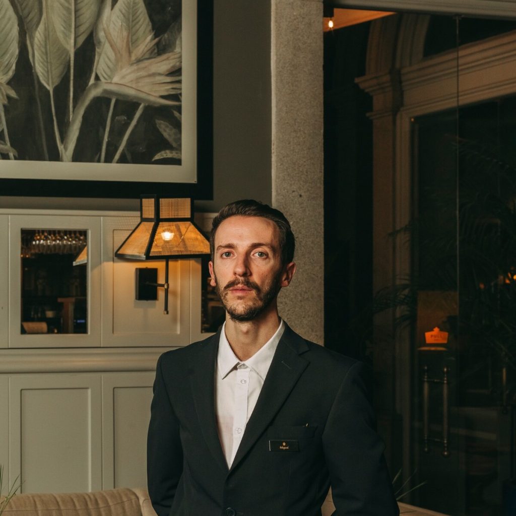 Man in a suit standing indoors near a wall with framed artwork.