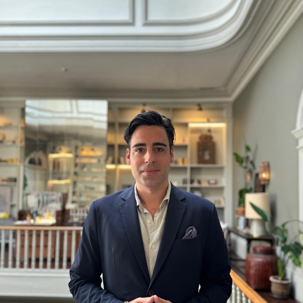 Man in a suit in a stylish room with shelves and decor in the background.