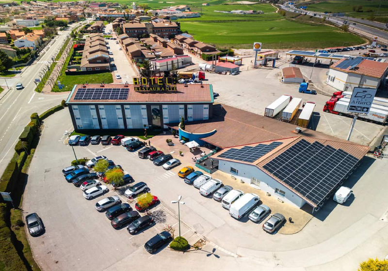 Vista aérea de un hotel y restaurante con estacionamiento, rodeados de casas y un campo verde en el fondo.