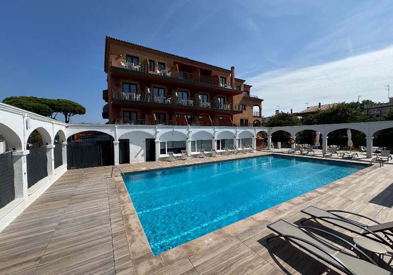 Piscina al aire libre frente a edificio de varios pisos, rodeada de tumbonas y arcos blancos, cielo despejado.