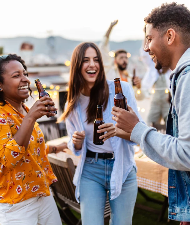 Personas riendo y brindando con botellas en una fiesta al aire libre con luces decorativas.