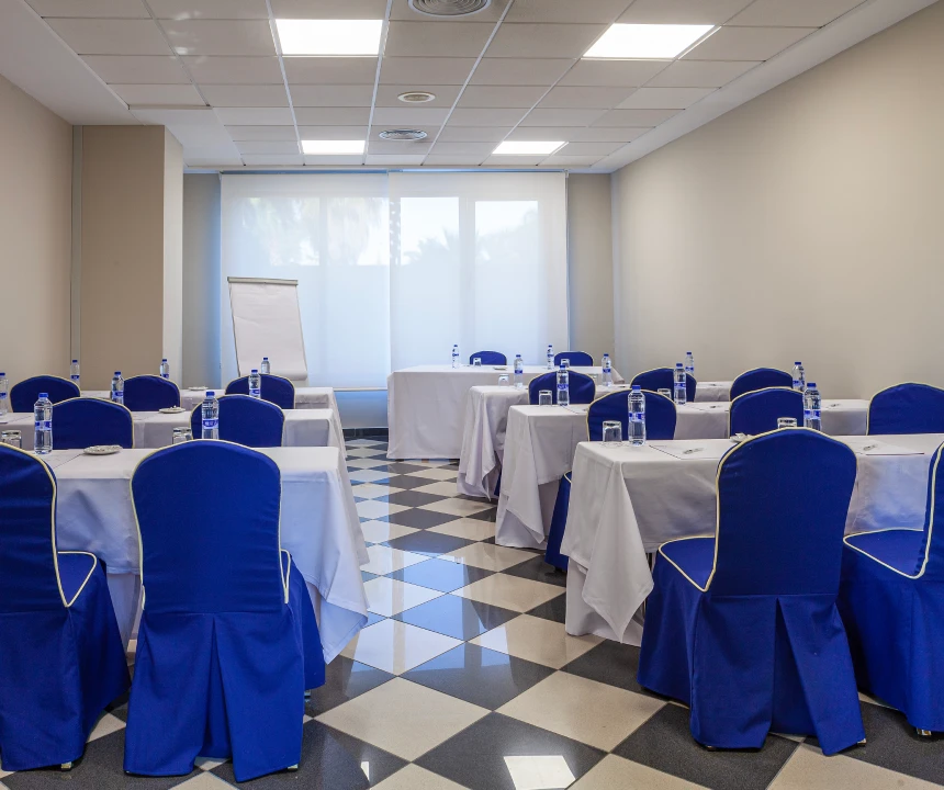 Sala de conferencias con sillas azules, mesas blancas, agua y un rotafolio frente a la ventana.