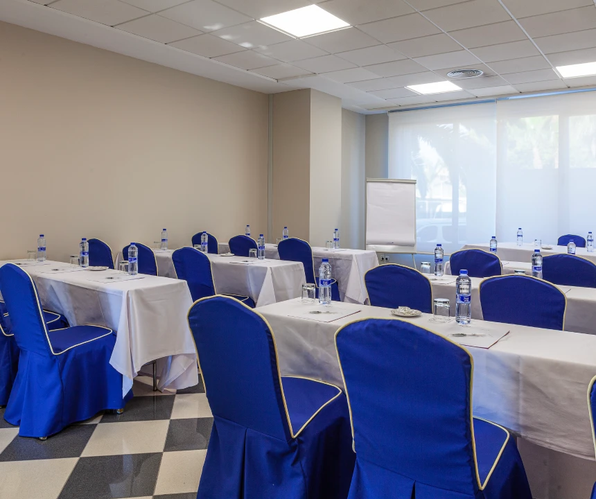 Sala de conferencias con mesas blancas y sillas azules, preparada con botellas de agua.