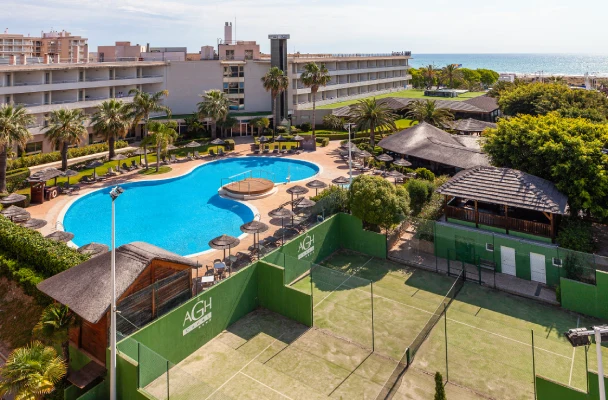 Hotel con piscina y canchas de tenis cerca del mar, rodeado de jardines y palmeras.