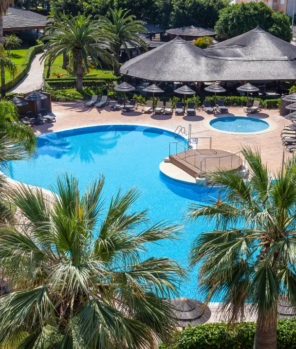 Piscina rodeada de palmeras y sombrillas de paja, con c&eacute;sped verde y sendero cercano.