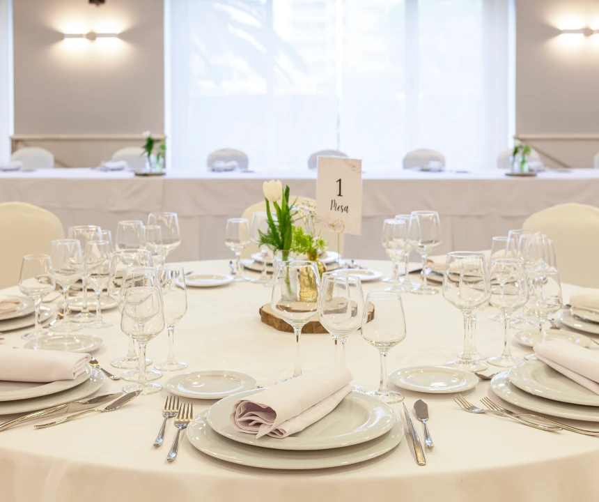 Mesa elegante con platos, copas, cubiertos y centro de mesa floral en un sal&oacute;n iluminado.
