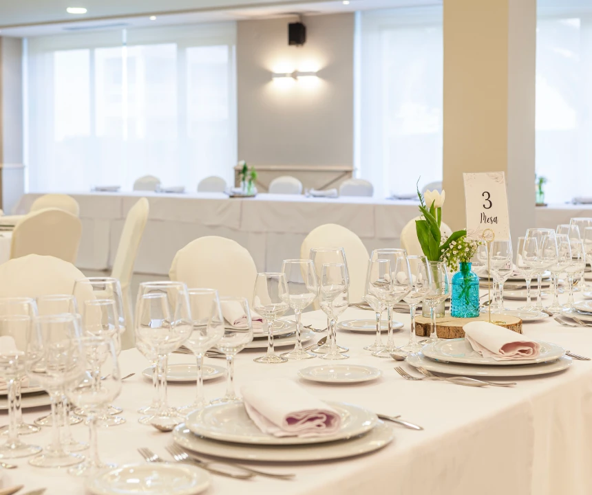 Mesa elegante con cubiertos, copas y flores en un sal&oacute;n iluminado.