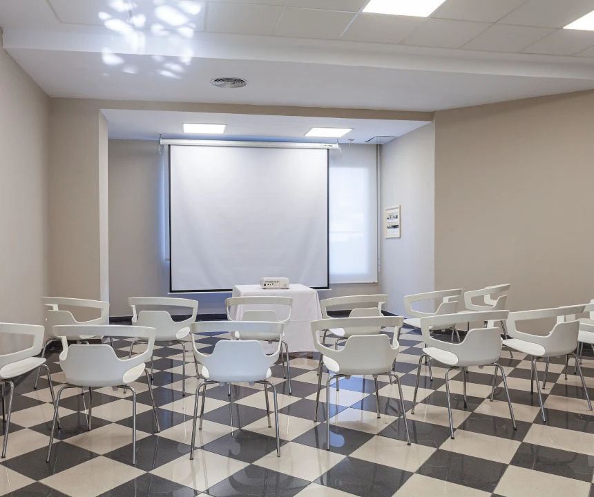 Sala de conferencias con proyector, pantalla y sillas blancas en suelo a cuadros.