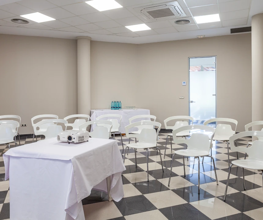 Sala de conferencias vac&iacute;a con sillas blancas y mesa con proyector. Suelo de baldosas blancas y negras.