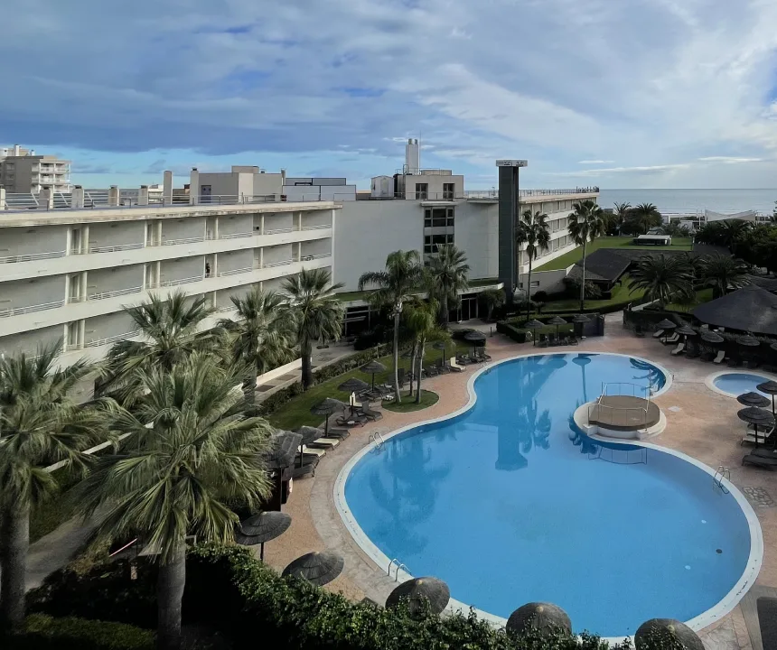 Hotel con piscina ovalada rodeada de palmeras, edificio moderno y cielo parcialmente nublado.