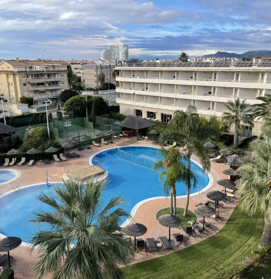 Vista a&eacute;rea de una piscina en un complejo con sombrillas, palmeras y edificios alrededor.