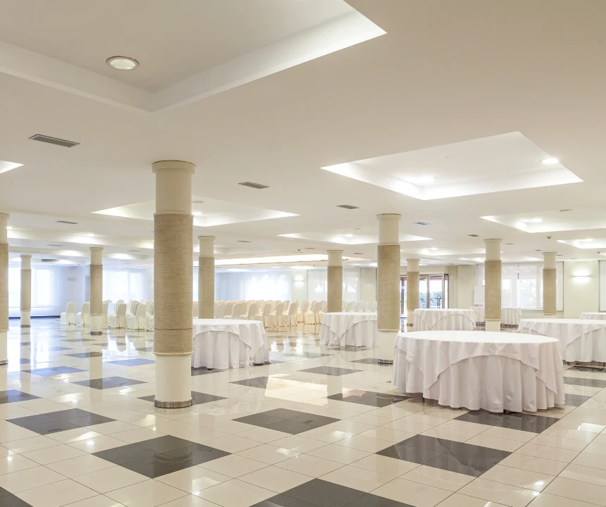 Sal&oacute;n de eventos vac&iacute;o con columnas, mesas redondas y sillas blancas. Piso de mosaicos.
