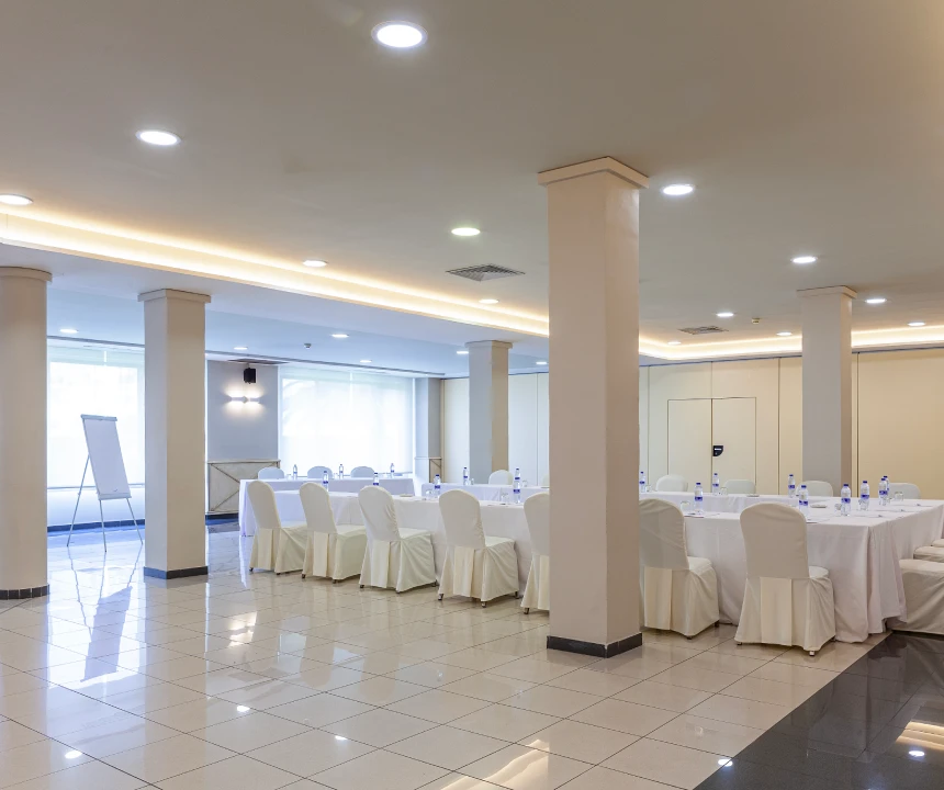 Sala de conferencias con mesa grande y sillas blancas, pizarr&oacute;n y botellas de agua.