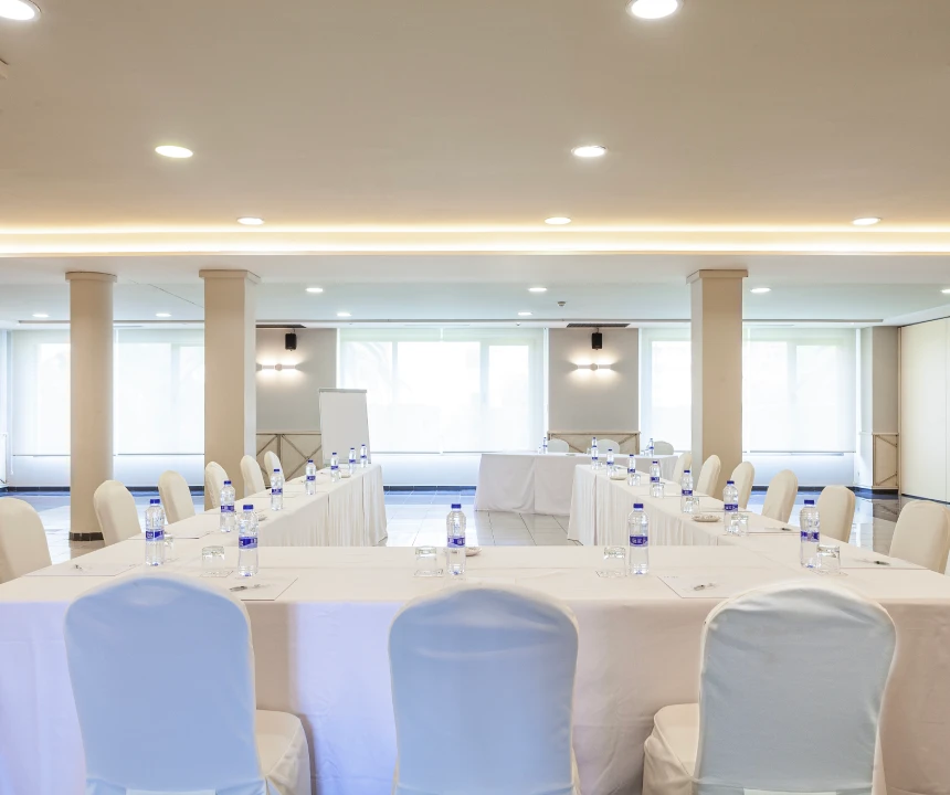 Sala de conferencias vac&iacute;a con mesas en forma de U, sillas blancas y botellas de agua.