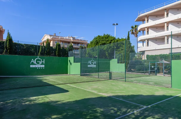 Cancha de tenis al aire libre con vallas verdes, rodeada de edificios y &aacute;rboles.