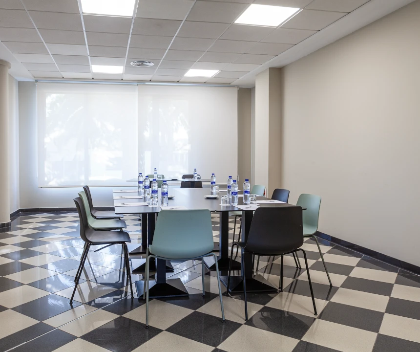 Sala de reuniones con mesa cuadrada, sillas y botellas de agua sobre un suelo a cuadros.