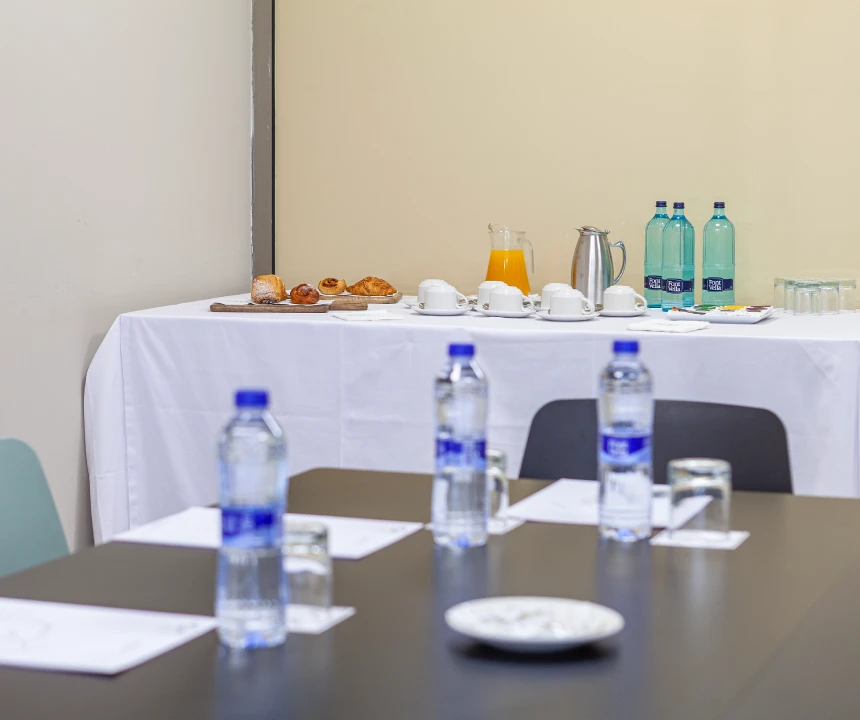 Mesa con agua y vasos; al fondo, mesa con caf&eacute;, jugo, pasteles y botellas de agua.