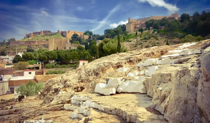Vista de un castillo en una colina, rodeado de vegetaci&oacute;n y rocas, bajo un cielo despejado.