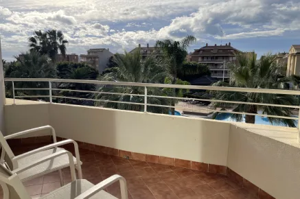 Balc&oacute;n con sillas, vista de palmeras y edificios, cielo parcialmente nublado.