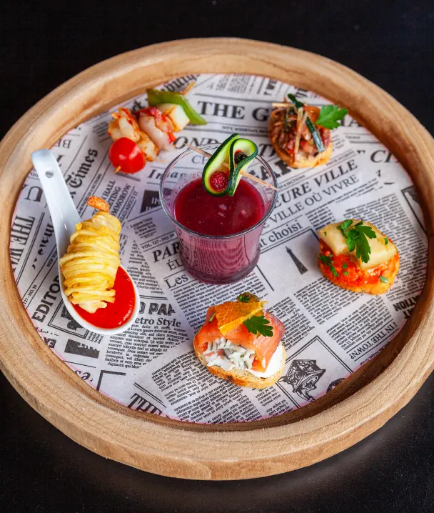 Una bandeja de madera con canap&eacute;s variados y una bebida morada sobre papel estilo peri&oacute;dico.