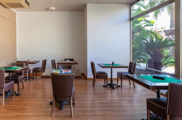 Sala de juegos con mesas y sillas marrones, junto a una ventana grande y plantas afuera.