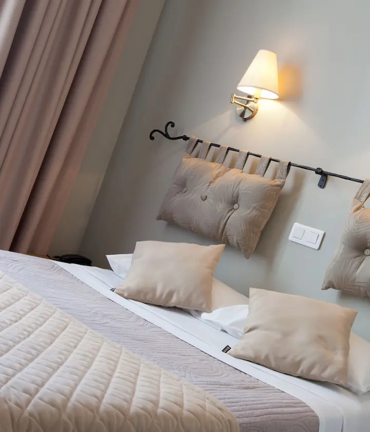 Lit avec coussins suspendus, lampe murale, rideaux et couvre-lit beige.