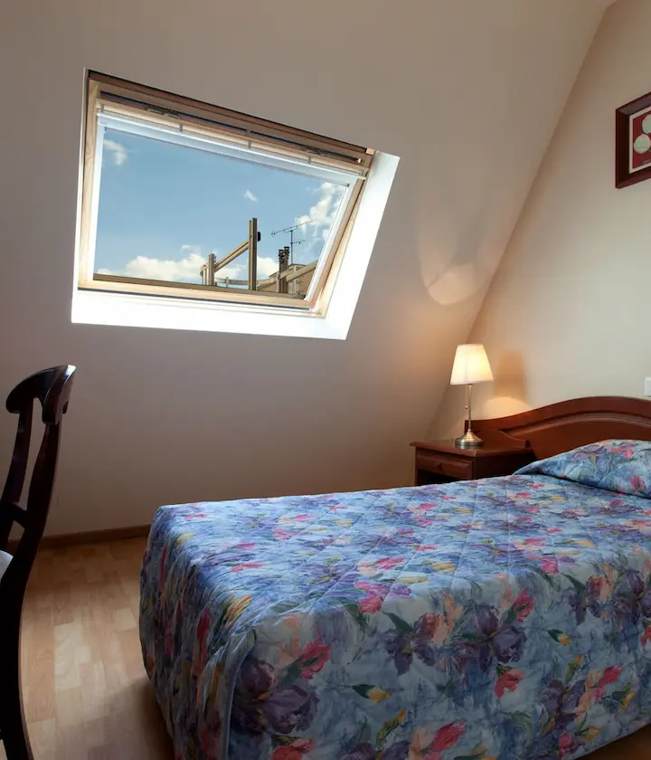 Chambre mansardée avec lit fleuri, fenêtre de toit ouverte sur ciel bleu et lampe de chevet.